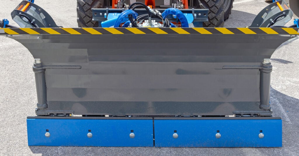 A truck in a parking lot is adjacent to a rolling truck. There is a snow plow blade attachment in front of the vehicle.