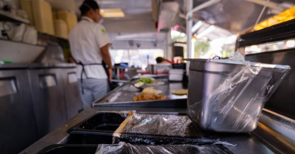 The interior of a food truck. A chef stands, facing the truck's cab. Food is on the grill and in plastic-wrapped containers.