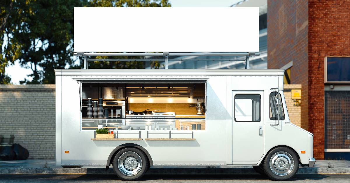 A white food truck parked along a street in front of a cement wall and adjacent to a red brick building.