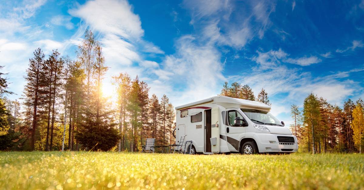 An RV parked in a grassy field with a forest behind it. The sun beams down, bathing the grass in light.