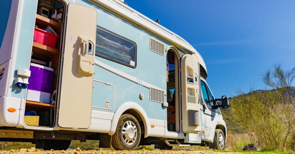 An RV parked on a dirt gravel road. The side storage panel is open, and various storage bins are on shelves.