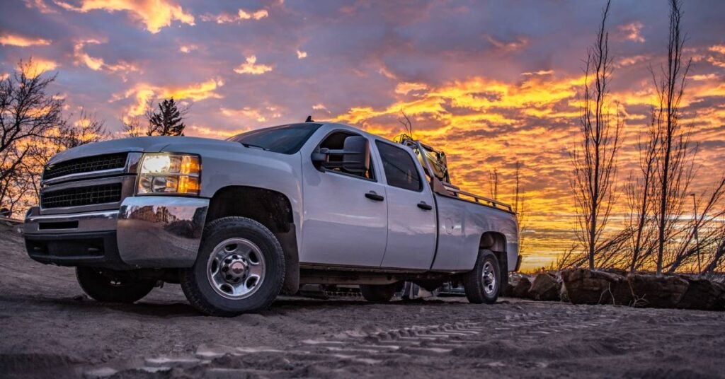 A white work truck sits on a gravel road at sunset. Dead trees and clouds paint the background of the image.