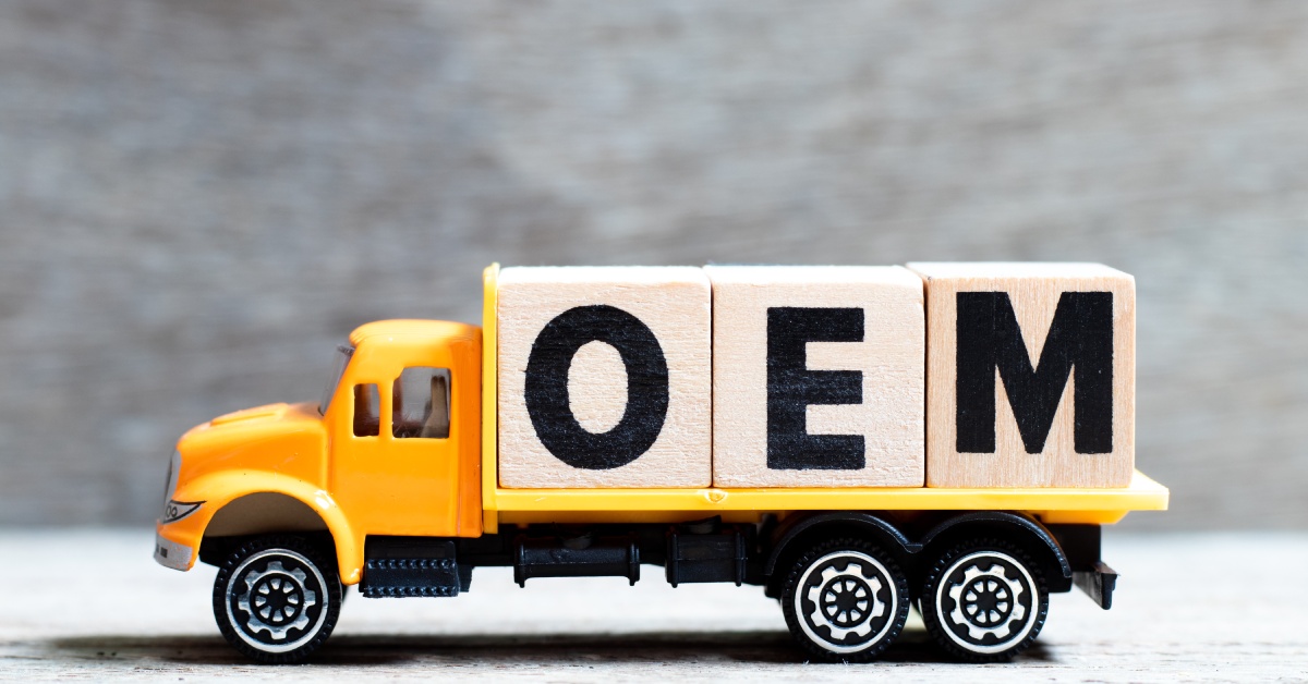 A yellow model truck holding letter blocks that spell "OEM." The truck has shiny wheels and is resting on a white surface.