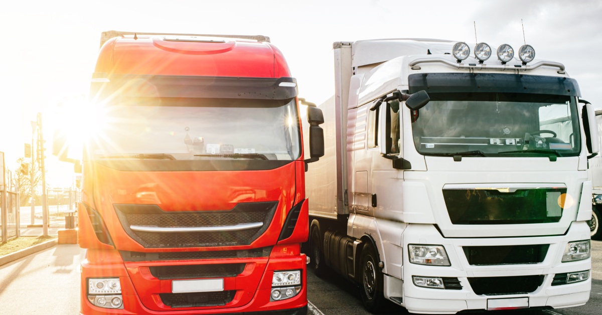 Two large trucks, one red and the other white, sitting in their parking spaces. The sun is shining behind them.