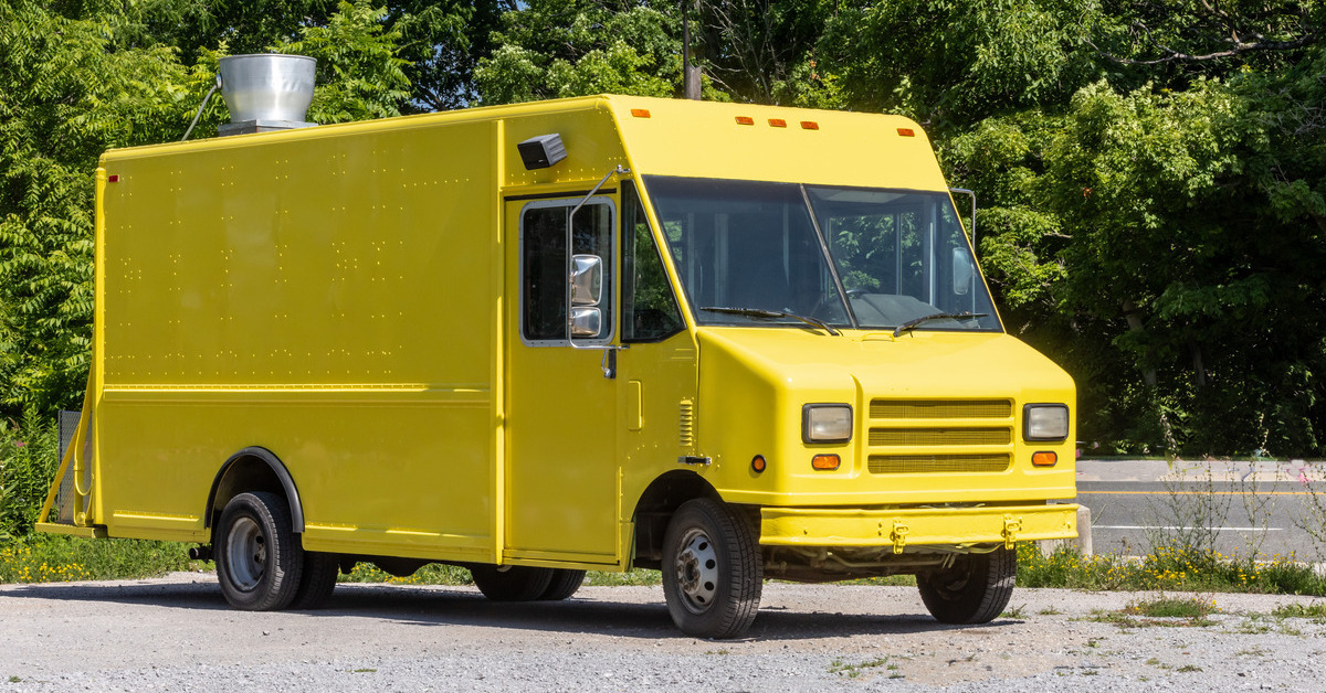 A yellow multi-stop truck sits in a rocky parking lot. Behind the truck are trees and a highway with a yellow line.