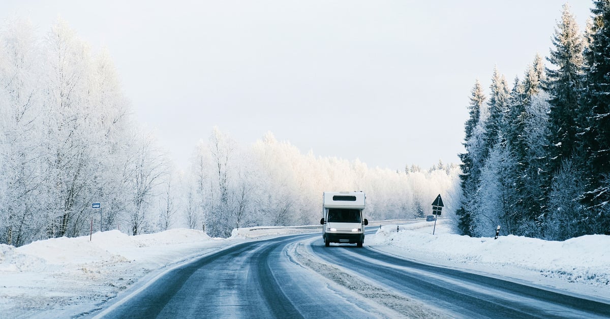 An RV drives on a long, winding road in winter. Snow is visible on the trees and the side of the road.