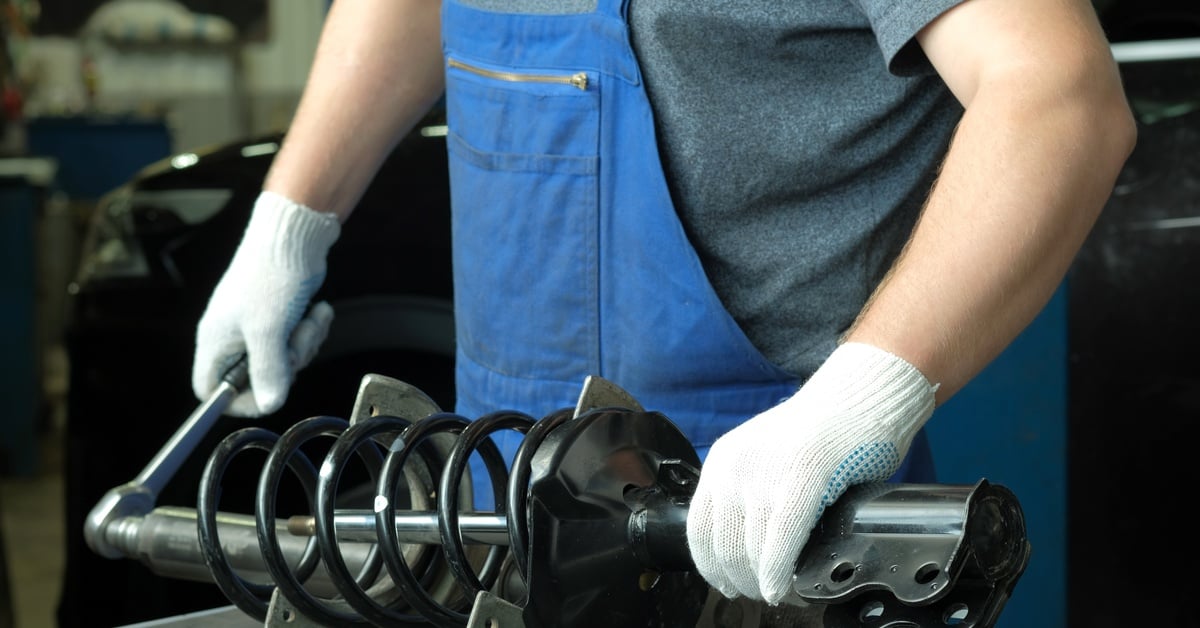 A man wearing blue overalls, a grey t-shirt, and white gloves handles a vehicle's suspension, including the strut and shocks.