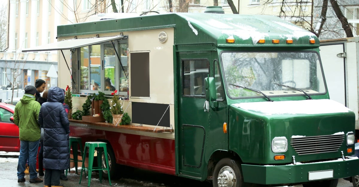 A green food truck stands on a city street with Christmas decorations on the windowsill. People wait outside for food.