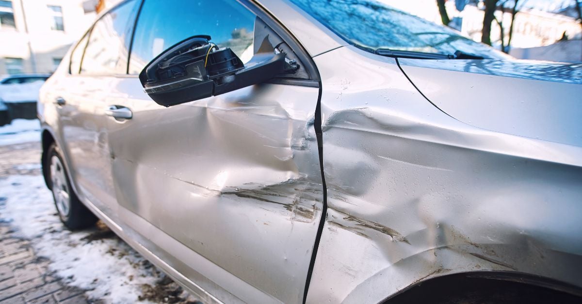 A close-up of a dented passenger door, side mirror, and front body panel on a silver sedan in a snowy driveway.