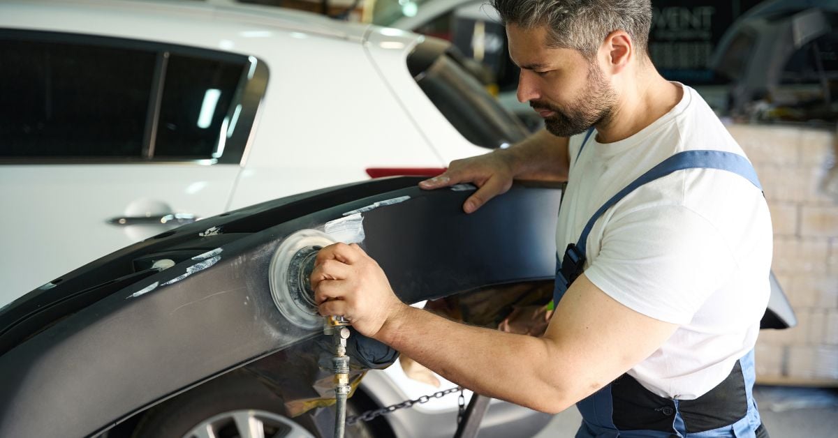 A repair technician in a body shop holds a silver car bumper while using a powered buffer to polish it.