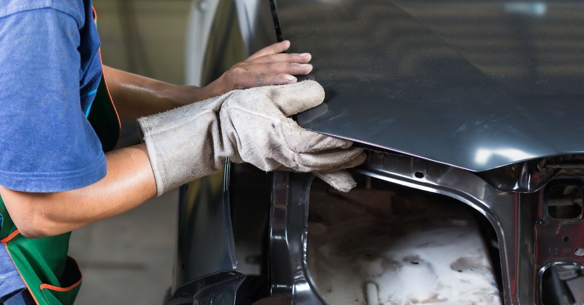 A repair technician wearing an apron and one work glove positions a replacement hood panel on a sedan.