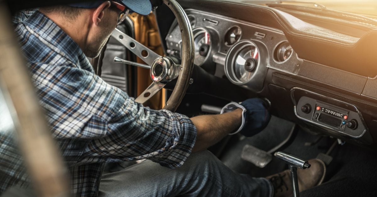 A mechanic wearing a plaid shirt and gloves sitting in the driver's seat of a classic muscle car, starting it up.
