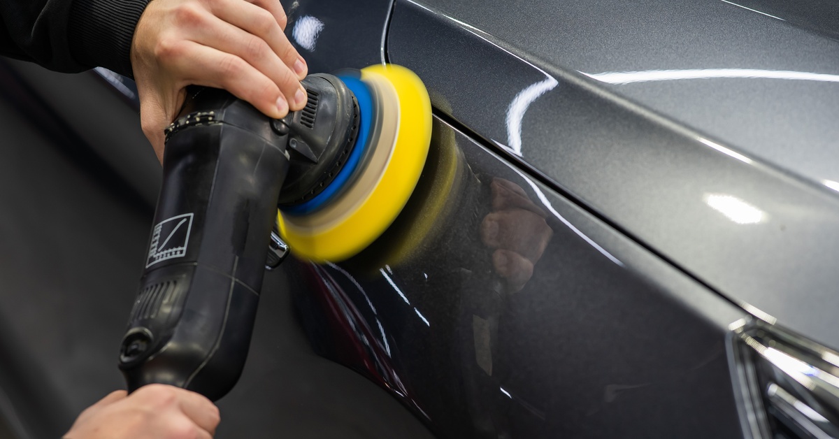 A close-up of a car detailer using a yellow handheld polishing tool on the fender of a dark gray vehicle.