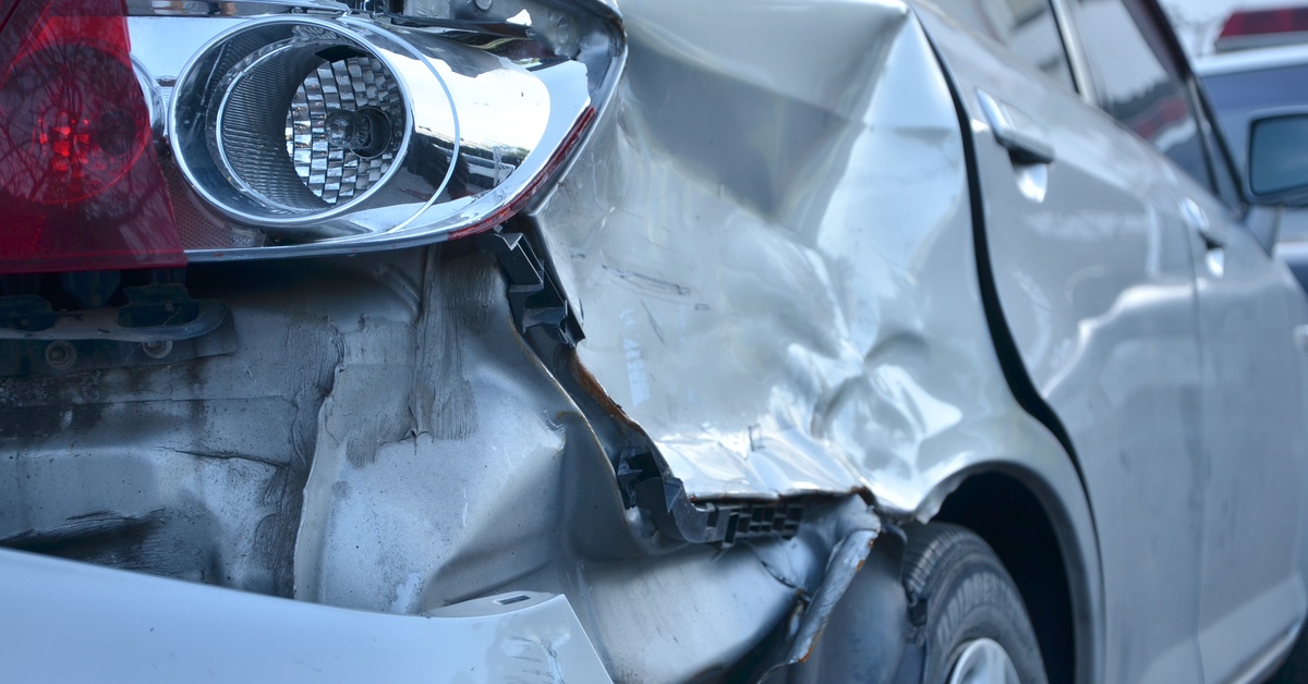 A close-up of a damaged white sedan bumper, which has become detached below the passenger side rear headlight.