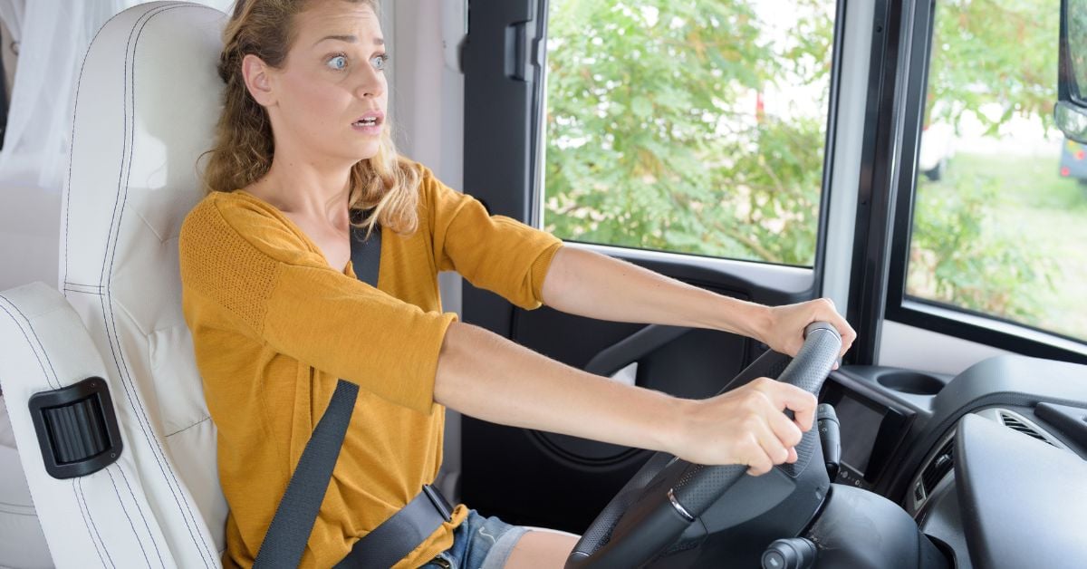 A woman in a yellow sweater, with a scared expression, is driving a camper van with white seats, gripping the wheel.