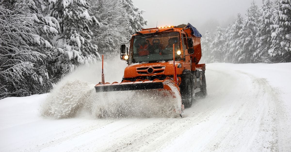 An orange snow plow removes snow from an icy road lined with snow-covered evergreen trees under an overcast sky.