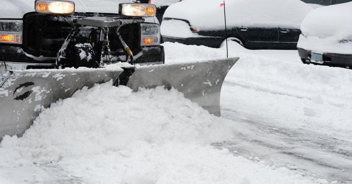 A snow plow attached to a white pick-up truck accumulates a small pile of snow while driving on a street.