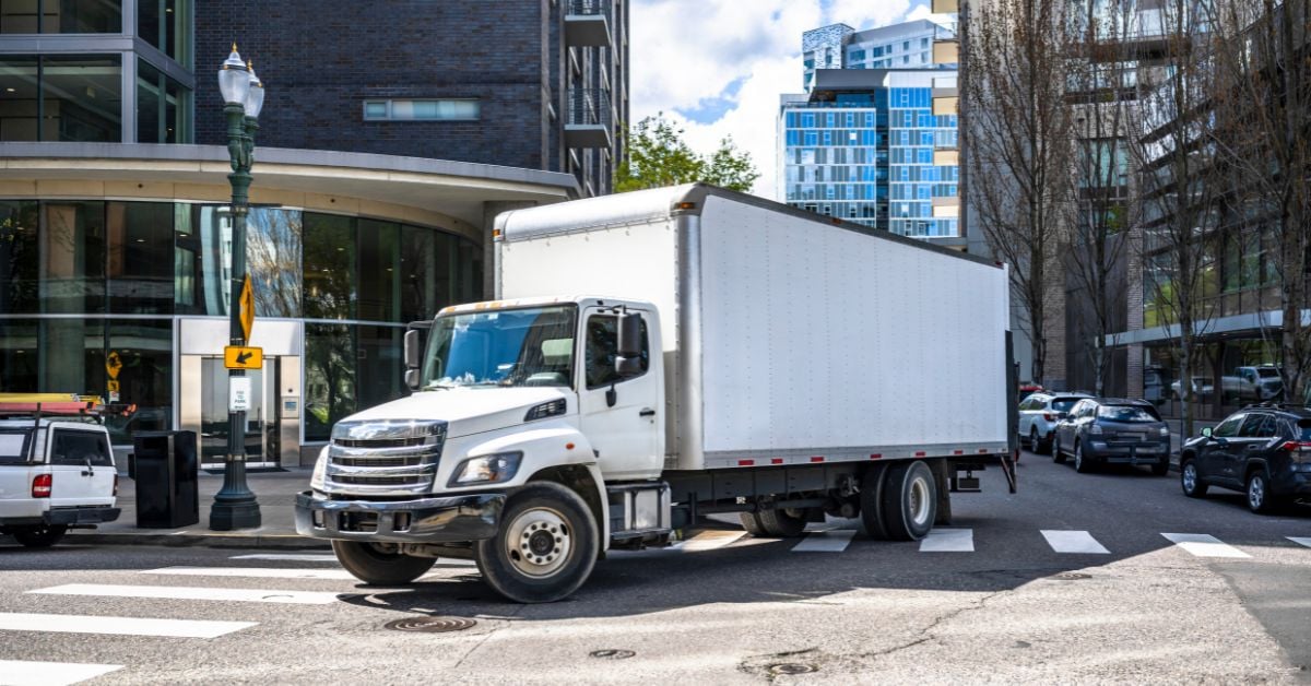 A white box truck is taking a right turn in an urban city with high-rise apartments and offices in the background.