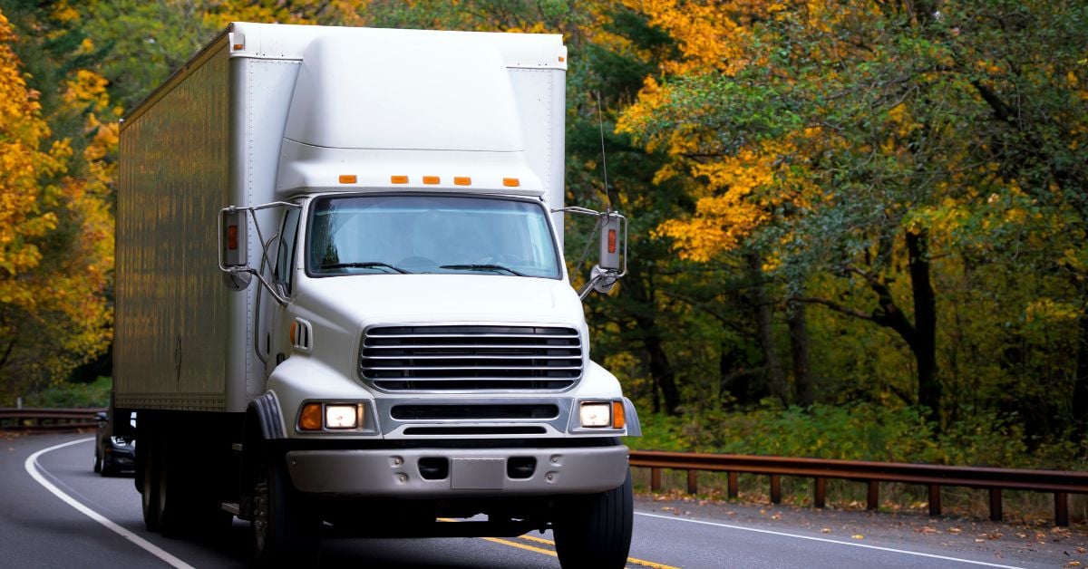 A white box truck is driving down a two-lane highway with green and yellow trees on each side, with a vehicle following the truck.