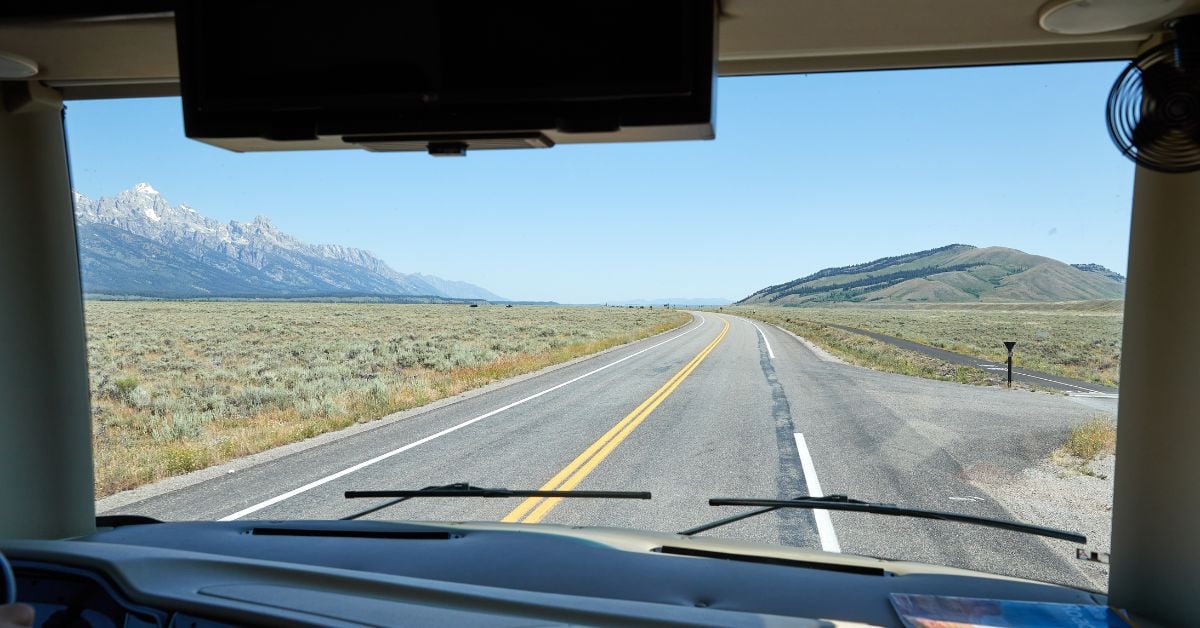 A long, open road curving left, leading toward a mountain range, seen from the interior cabin of an RV.