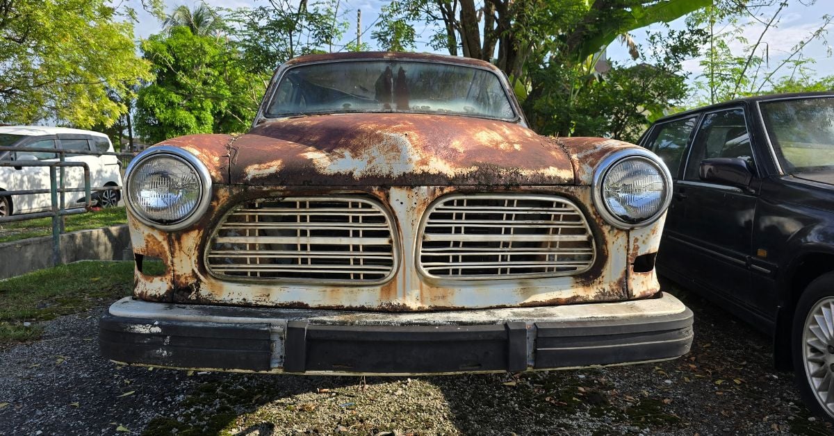 A classic vehicle with its hood covered in rust sits outside under a tree next to a dark blue vehicle.