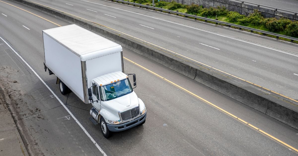 An aerial view of a white semi truck with a box trailer driving down an empty two-lane highway.