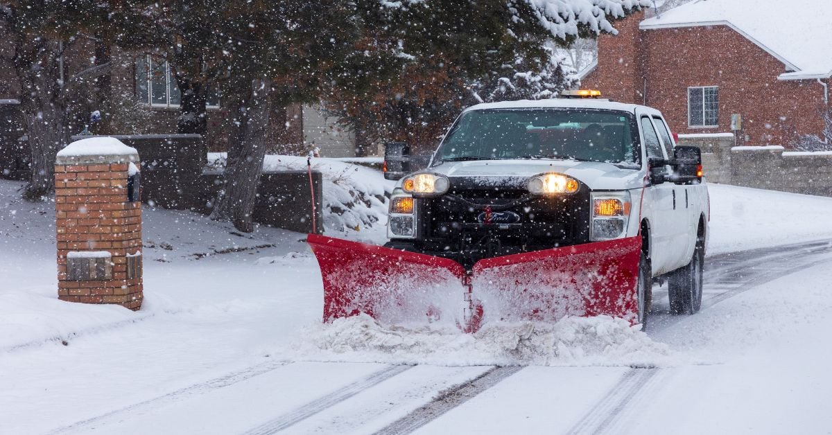 A white pick-up truck removes snow using a red V-shaped snow plow in a neighborhood with snow-covered brick houses.