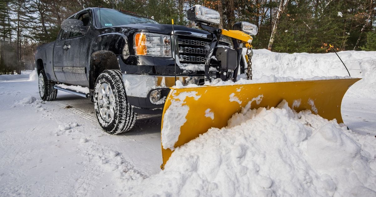A black pick-up truck with a mounted yellow straight-blade snow plow removes snow from a street lined with trees.