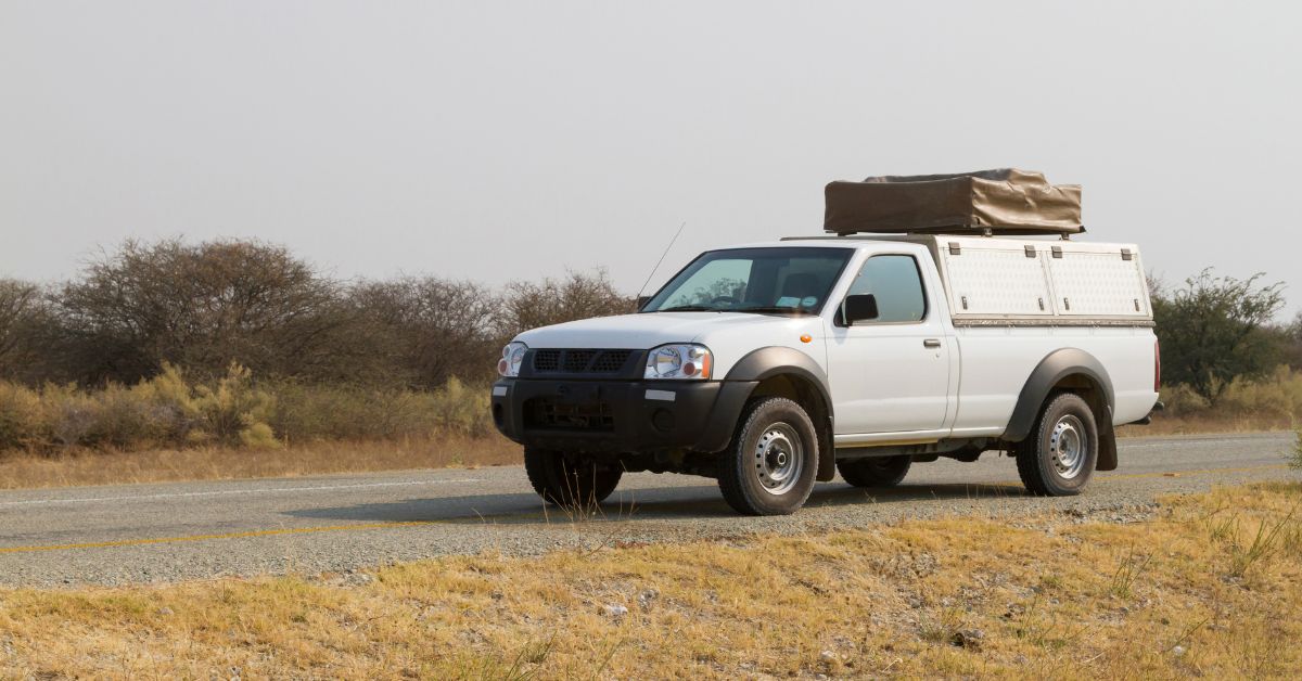 A white pickup truck with a covered bed is parked on the shoulder of an empty road in an arid climate.