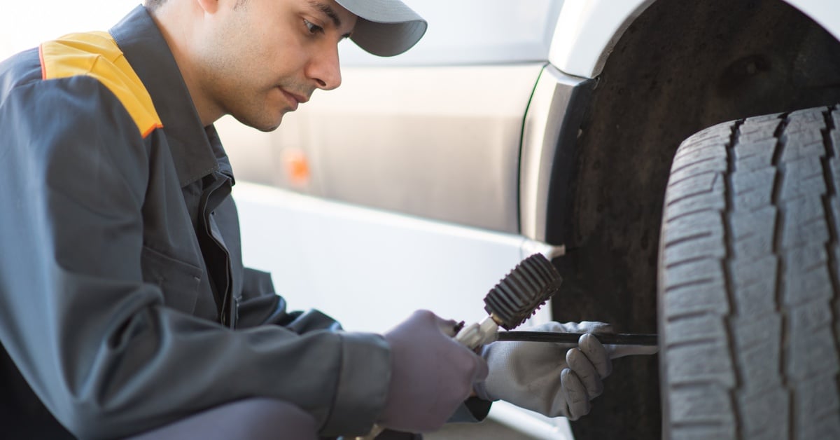 A male mechanic wearing coveralls and gloves uses a gauge to measure pressure on a van's front tire.