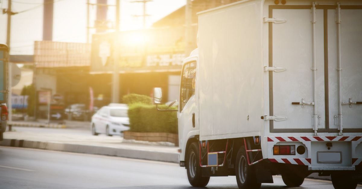 A white compact semi-truck drives on the road as the blinding sun shines on it. On the other side of the road is a taxi.