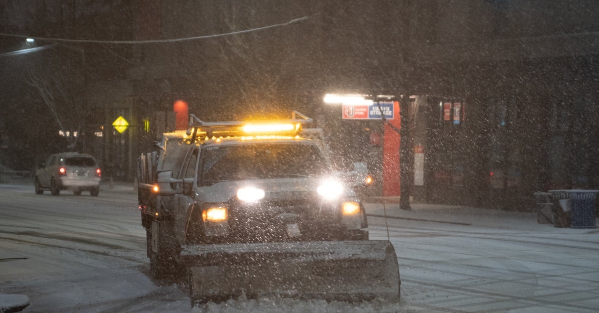 A white pickup truck with a snow plow blade drives on a street in low light, with its headlights and emergency lights shining.