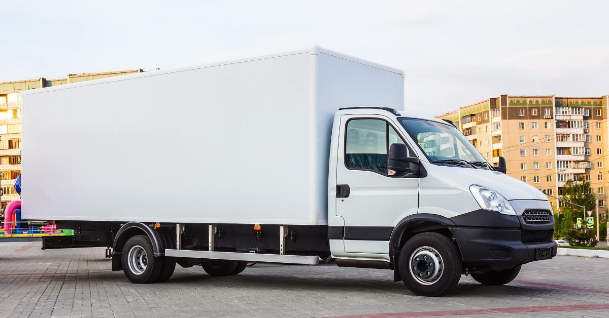A white commercial box truck is parked on gray and red stone bricks, in the middle of a residential area.
