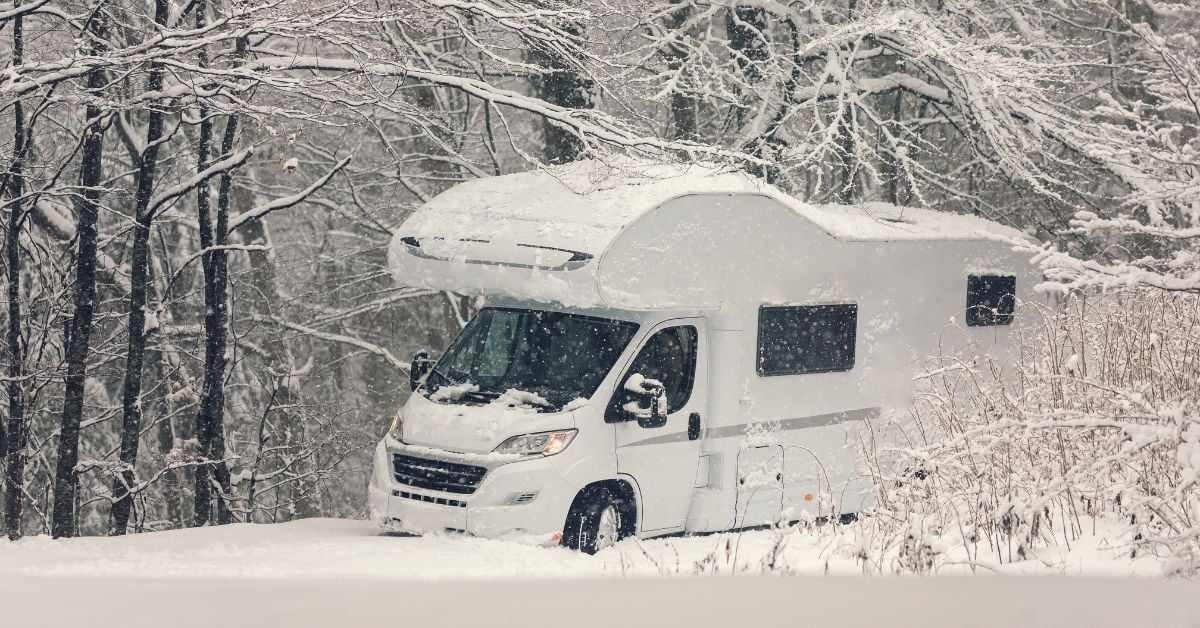A white RV is in a snow-covered forest with bare trees and bushes. The vehicle is also covered with snow.