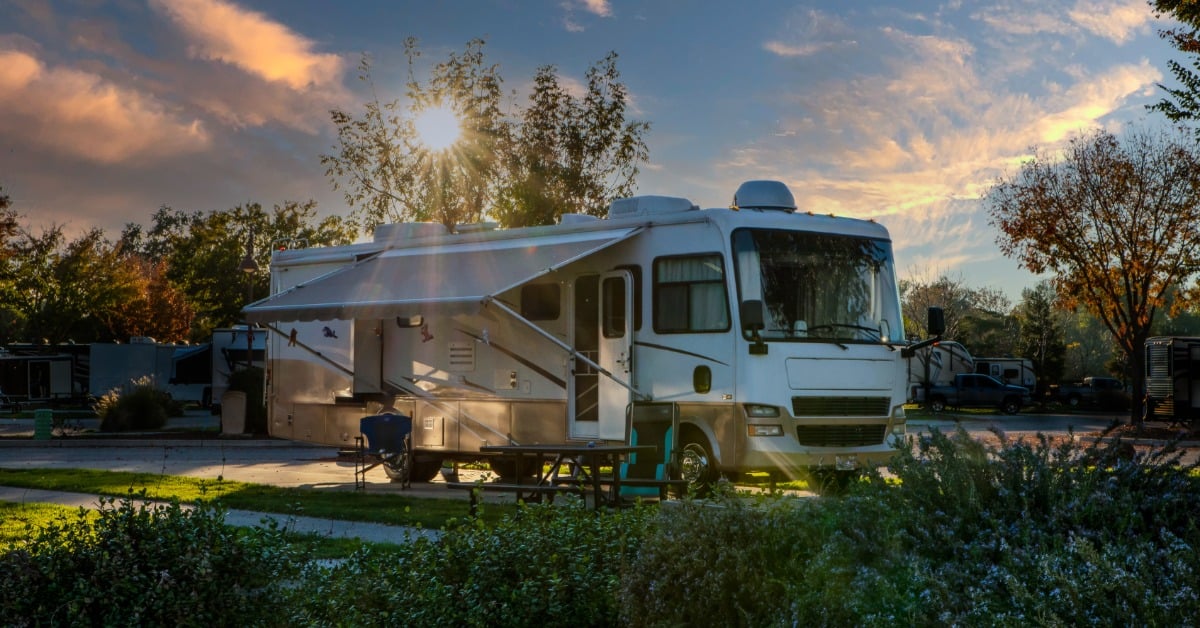 The sun peeks through the tree and shines down on a white, open RV parked on the road while surrounded by greenery.