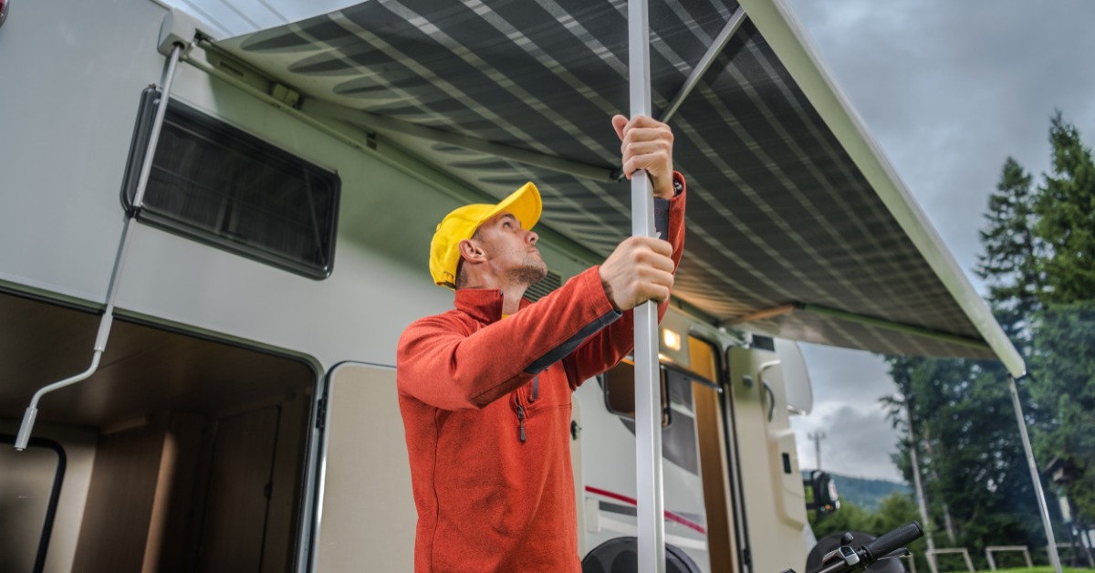 A man wearing a yellow hat and red jacket holds the arms of an RV's awning system as storm clouds roll in.