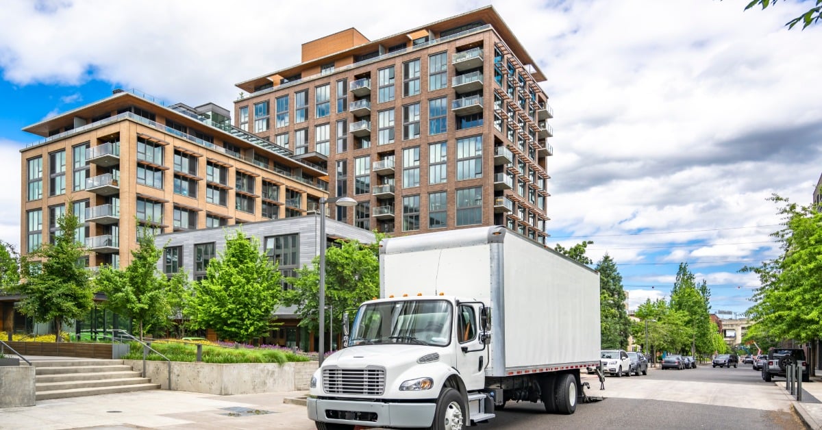 A white semi-truck with a box trailer load drives through the city street, surrounded by high-rise buildings.