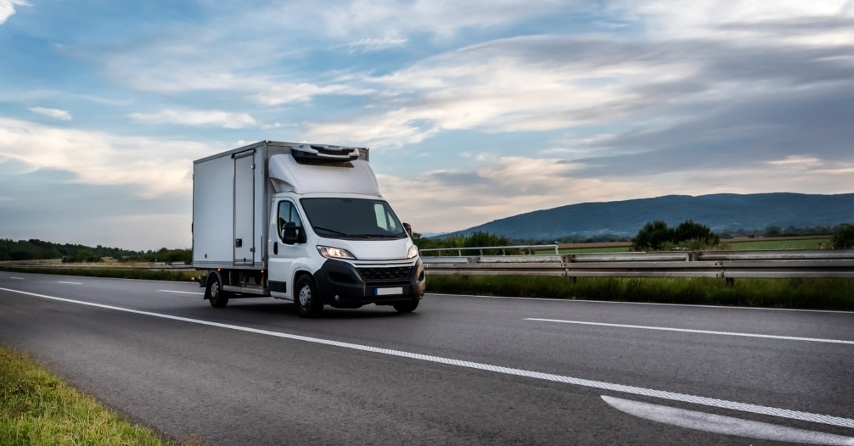 A white cargo van drives on a road, surrounded by grasslands. In the distance are a view of the mountains.