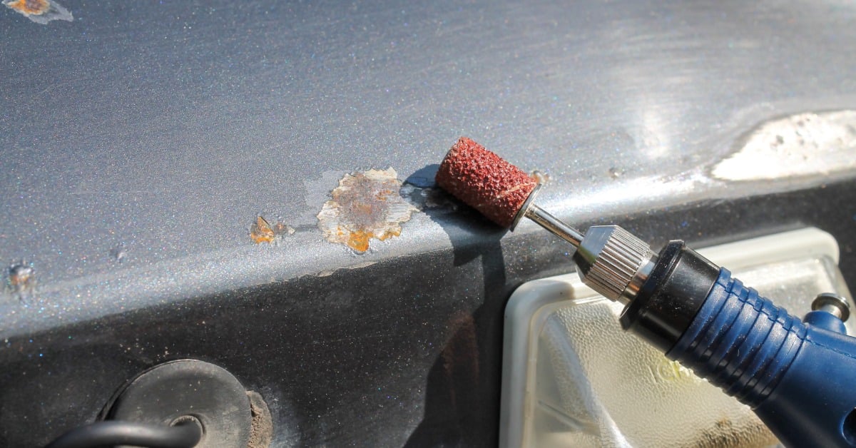 A person uses a blue and red rotary tool to scrap off the rust eating the metal on a dark gray vehicle.