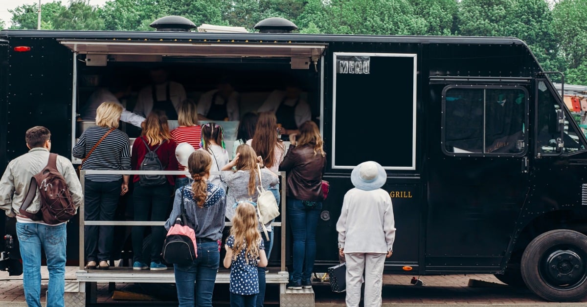A crowd of people stands in front of a black step van set up as a food truck, parked on red brick road.