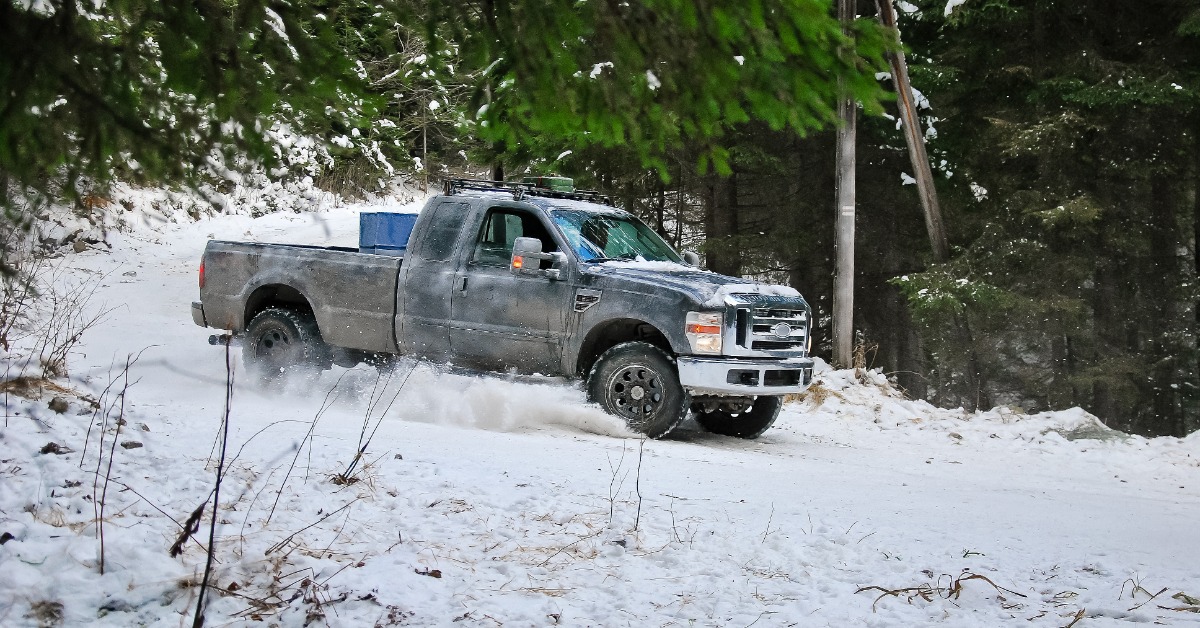 A black pickup truck drives through a pine forest filled with snow, kicking up snow and debris with its tires.