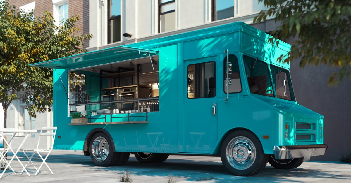 A bright aquamarine food truck parks in front of a residential building, next to a white table and a chair.
