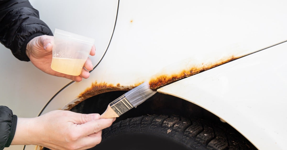 A person wearing a black puffer jacket uses a paintbrush to apply a rust encapsulator on a white vehicle.