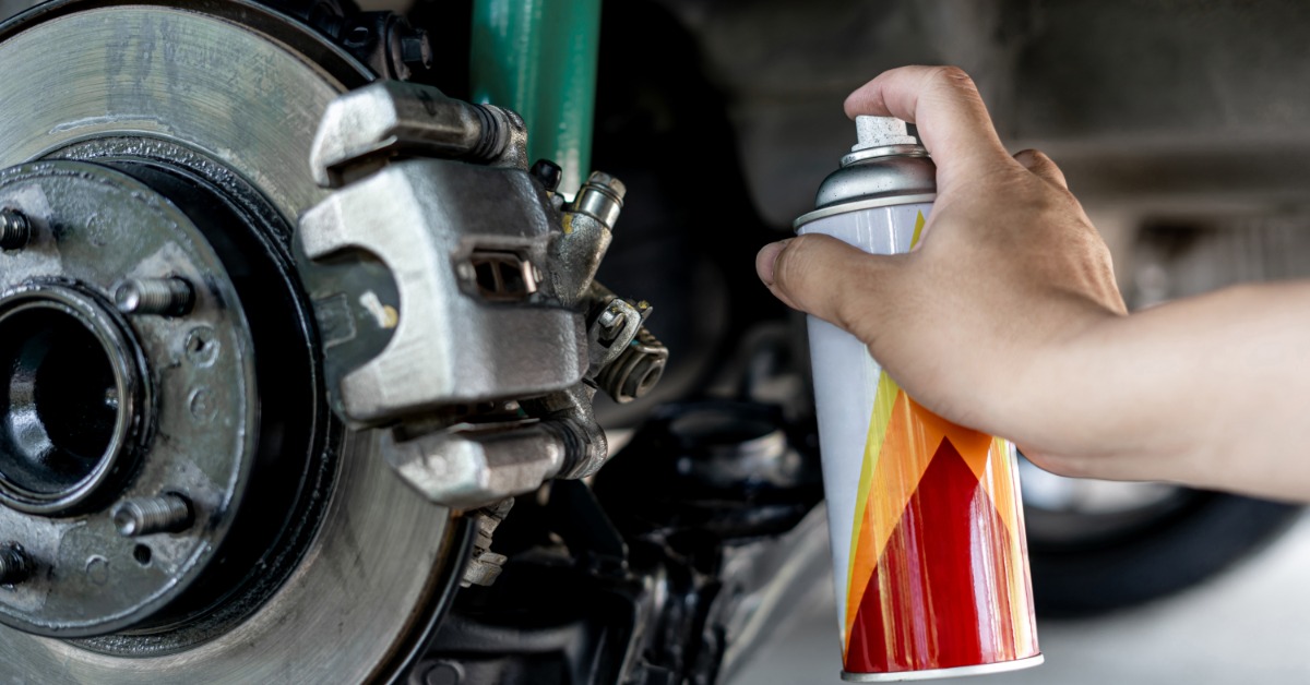 A person holding a rust protection spray can points it towards the brake plate of a lifted car's caliper.