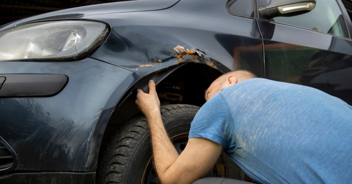 A man wearing a light blue shirt crouches down to inspect the rust damage on the fender of a black car.