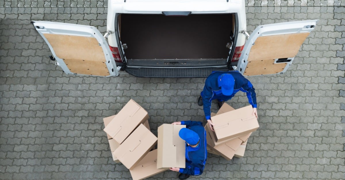 Two delivery workers wearing blue uniforms unloads stacks of cardboard boxes from a white van onto the ground.