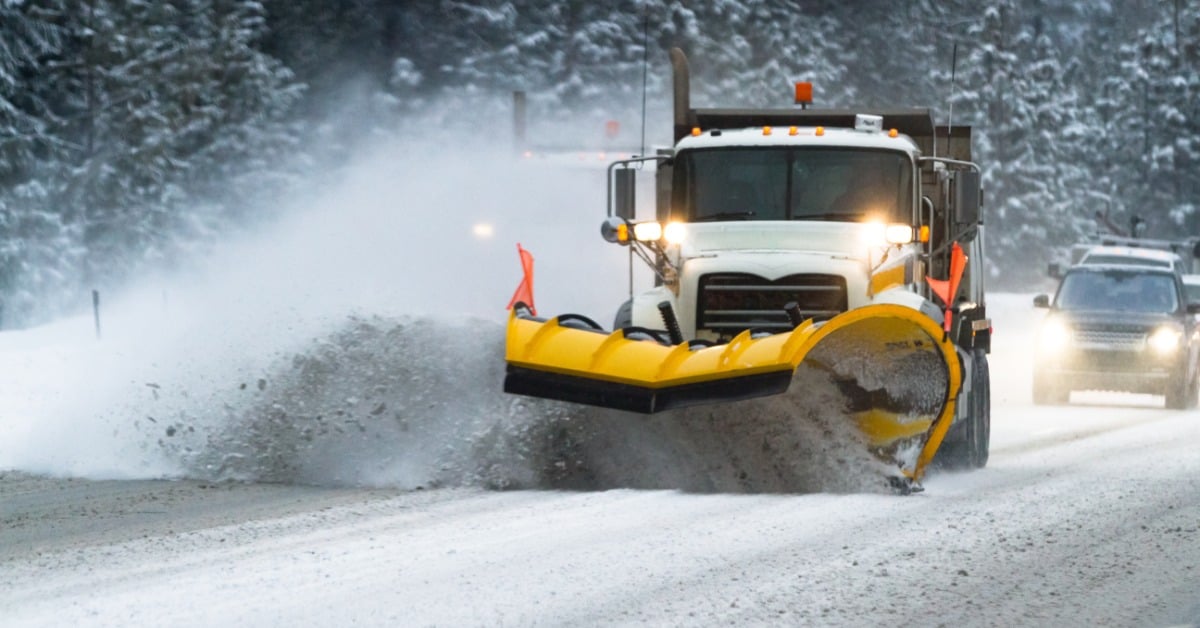 A white snow plow truck drives through the road, kicking up snow as it plows. Behind it are trees and cars.