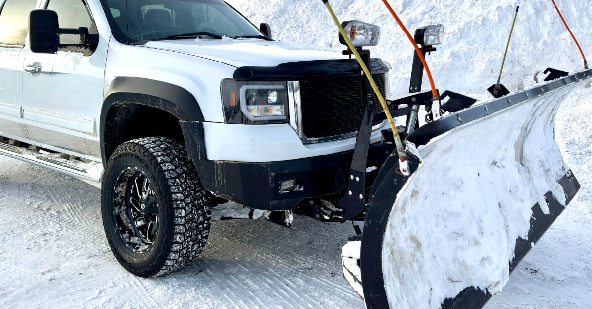 A white pickup truck equipped with a front snow plow, is currently parked and ready for winter storm cleanup.