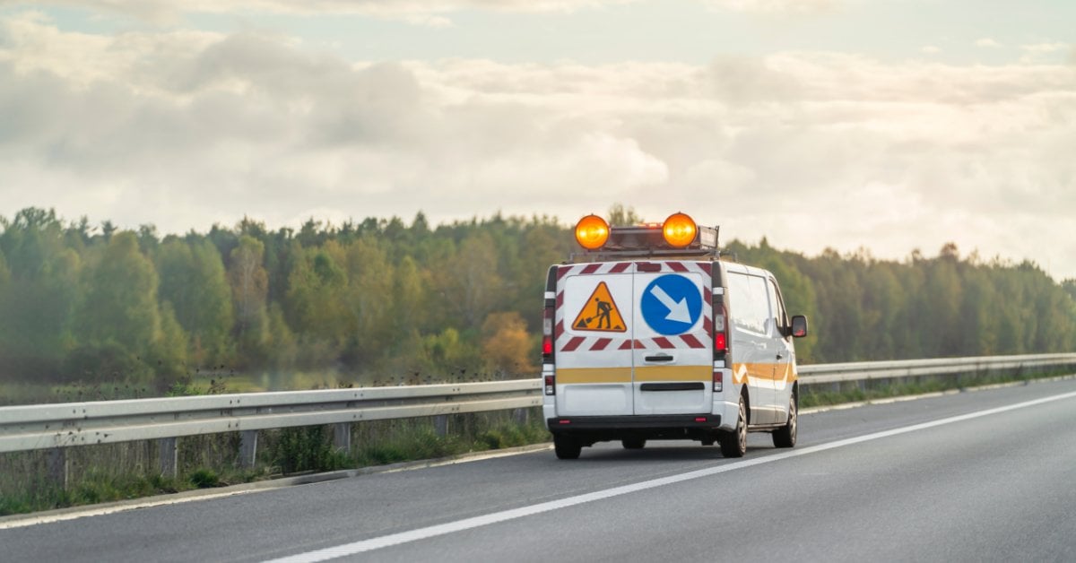 A highway patrol van drives on a roadway with amber roof beacons and warning signs on the rear doors.