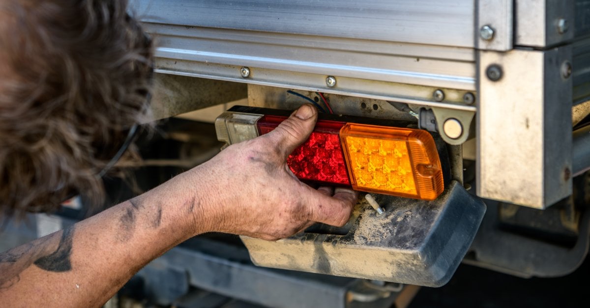 A mechanic holds and repairs the rear tail light on a utility vehicle truck, working near the lamp housing.